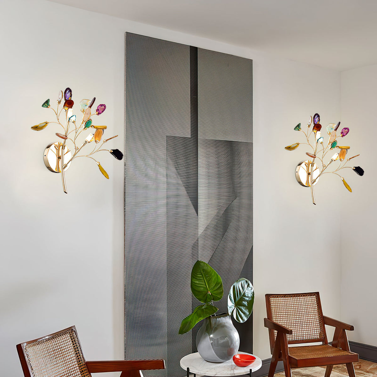 Two multicolour Clérachic agate wall lights on a white wall beside a grey panel, with cane chairs and a plant below.