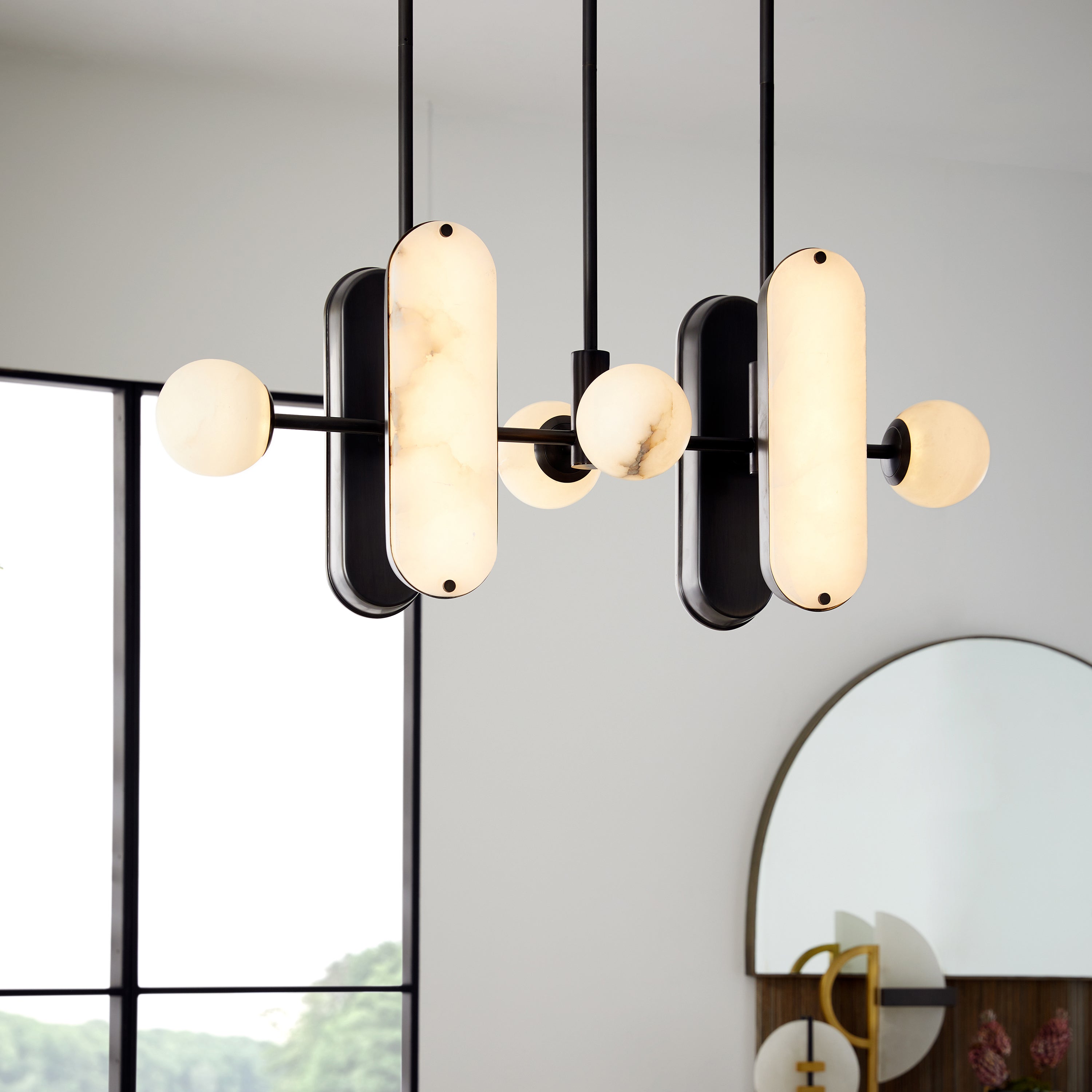The Clérachic Alabaster 5-Light Linear Island Pendant lit above a living room, glowing beside a black steel window and an arched mirror.