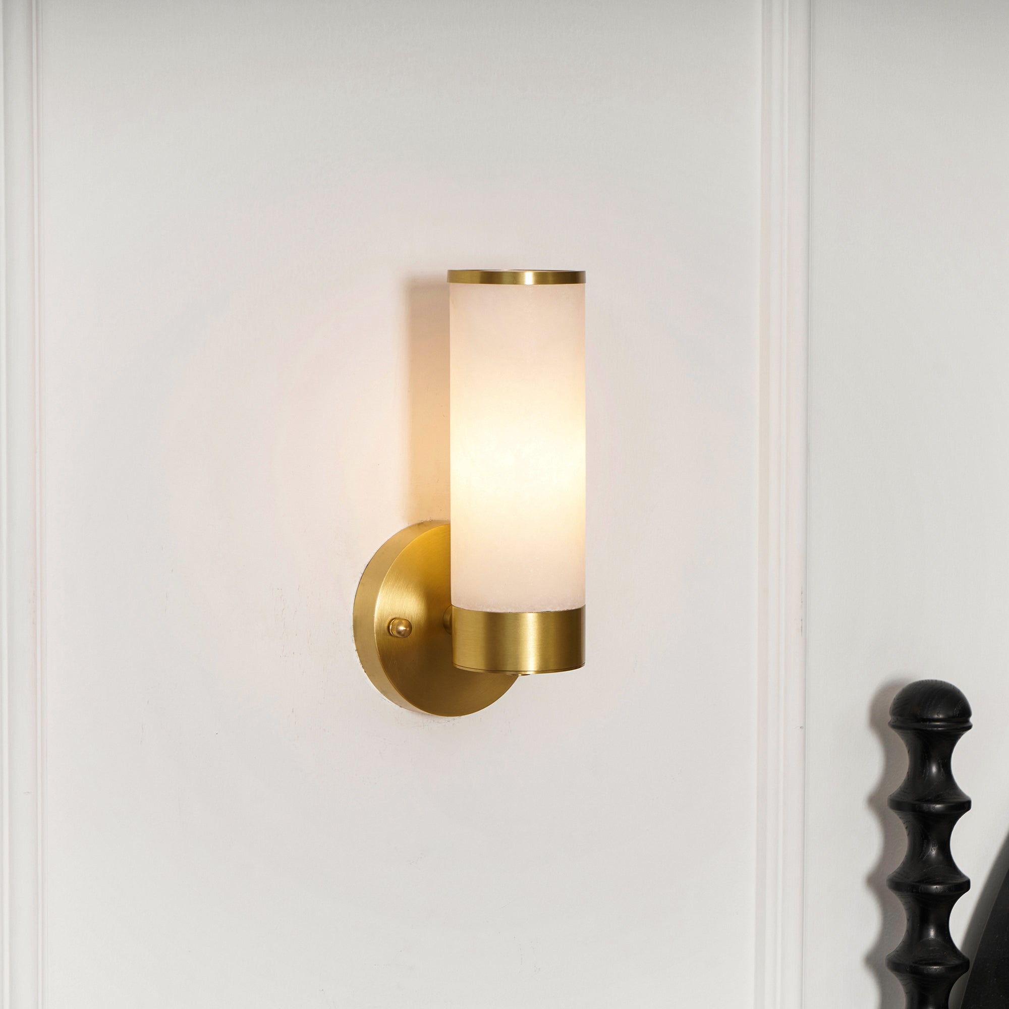 Clérachic Alabaster and Brass Wall Sconce lit on a panelled white wall, a soft halo beside a dark turned post.