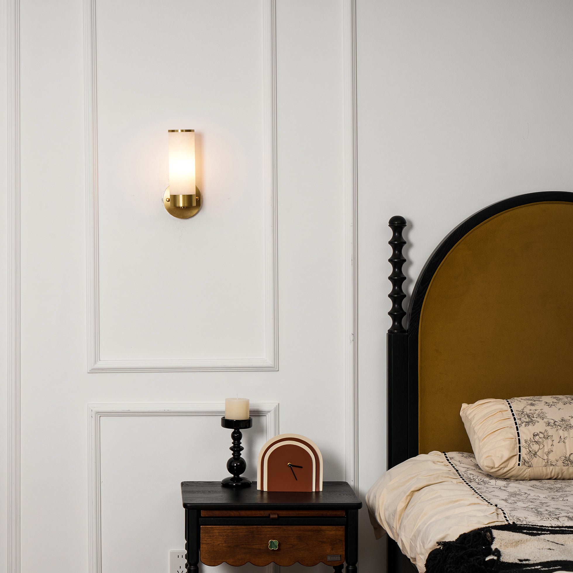 Clérachic Alabaster and Brass Wall Sconce lit beside a bed with an ochre headboard, above a black bedside table.