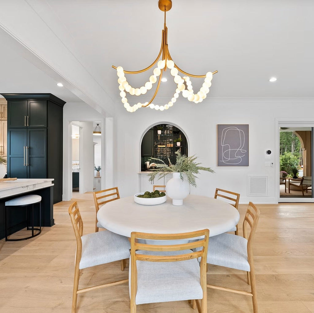 Clérachic Alabaster Bead Chandelier in brass above a round white dining table, lighting an open kitchen with dark cabinetry.