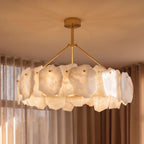 Clérachic round chandelier lit warm, its alabaster panels and brass arms and canopy seen from below in front of linen curtains.