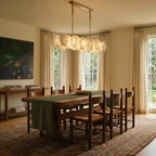 Clérachic linear alabaster chandelier over a long wooden dining table with rush-seat chairs and tall windows with linen curtains.