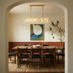 Clérachic linear alabaster chandelier framed by an arched doorway above a walnut dining table, with abstract art behind it.