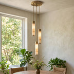 This Clérachic chandelier's brass-capped columns glow warm in a plastered corner beside a window and potted plants.