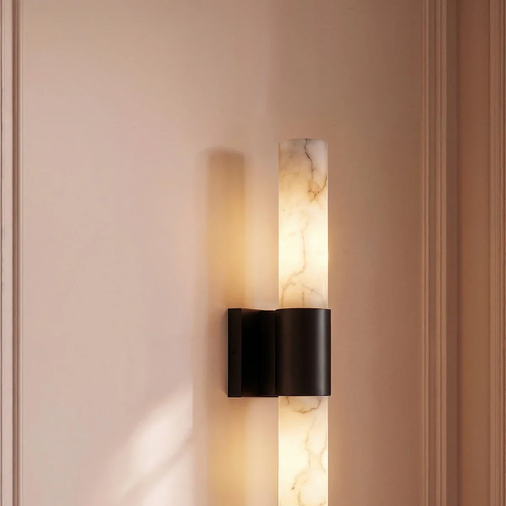 Clérachic wall sconce with a black band and glowing alabaster column on a blush panelled wall, its shadow cast beside it.