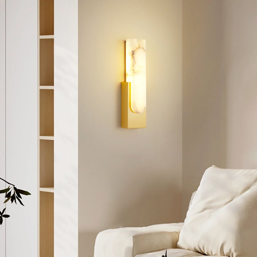 Lit Clérachic alabaster wall light with a gold-toned plate on a beige wall, beside a tall open shelf and the arm of a cream sofa.