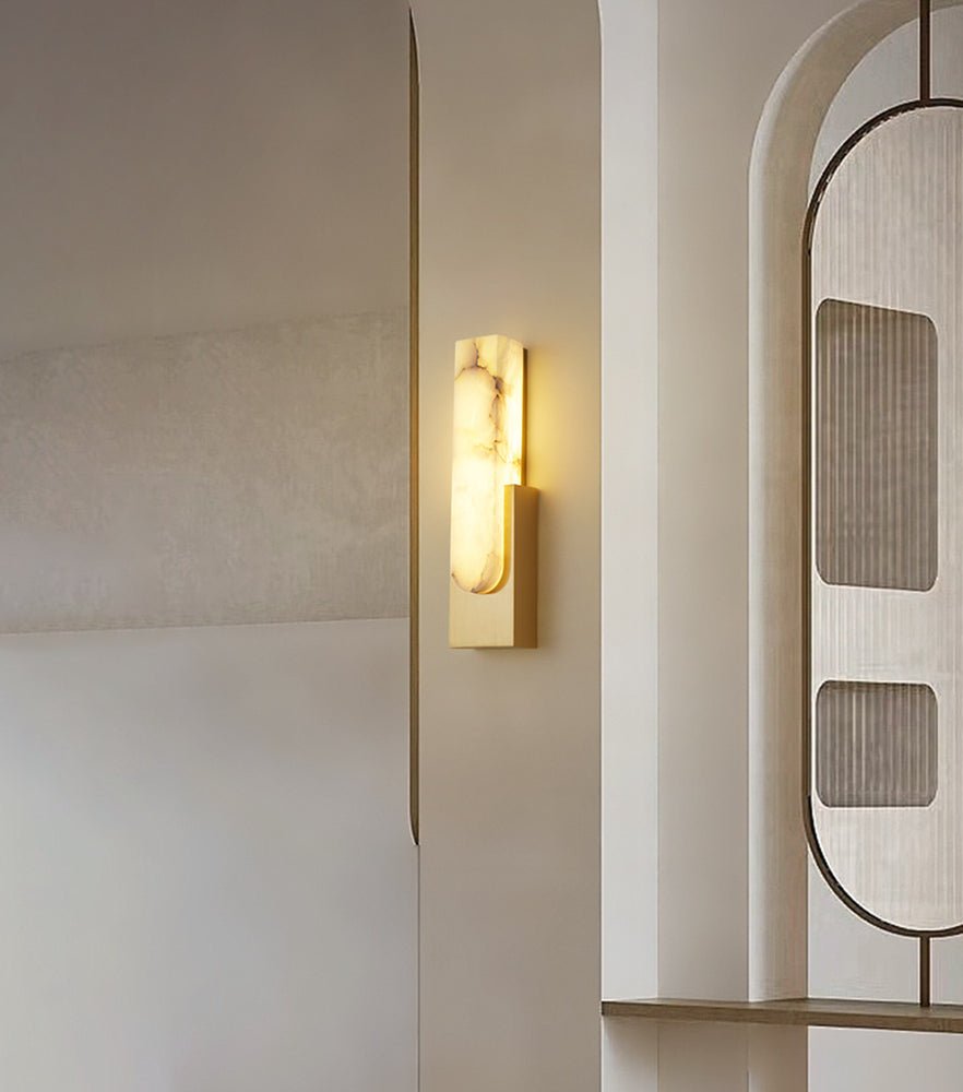 A gold-toned Clérachic wall light glowing beside an arched, glazed partition, its alabaster bar showing soft grey veining.