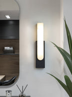 Black-backed Clérachic wall light with a white alabaster bar, lit, on a pale wall beside an oval mirror and a tall green leaf.