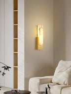 Lit Clérachic alabaster wall light on a beige wall beside a slim open shelf, a cream sofa and a pair of glasses on a table below.