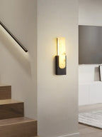 A black Clérachic wall light lit on a pale wall beside a staircase with a glowing handrail, a wall-mounted television behind.