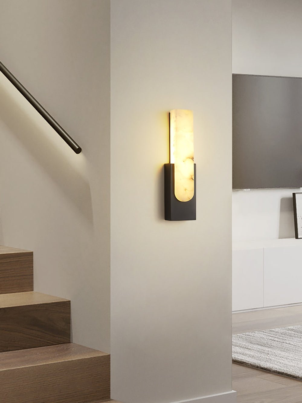 A black Clérachic wall light lit on a pale wall beside a staircase with a glowing handrail, a wall-mounted television behind.