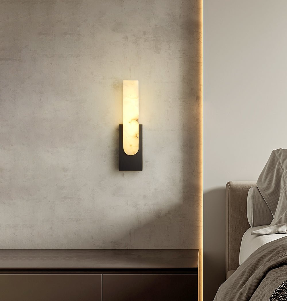 Lit Clérachic wall light with a black back plate on a stone-textured wall, above a dark sideboard and a bed with a grey throw.