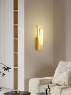Gold-toned Clérachic wall light lit on a cream wall, with a tall shelf at the left and a sofa cushion below.