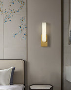 Gold-toned Clérachic wall light lit above a bedside table, on a panelled wall with a decorative blue and gold pattern.