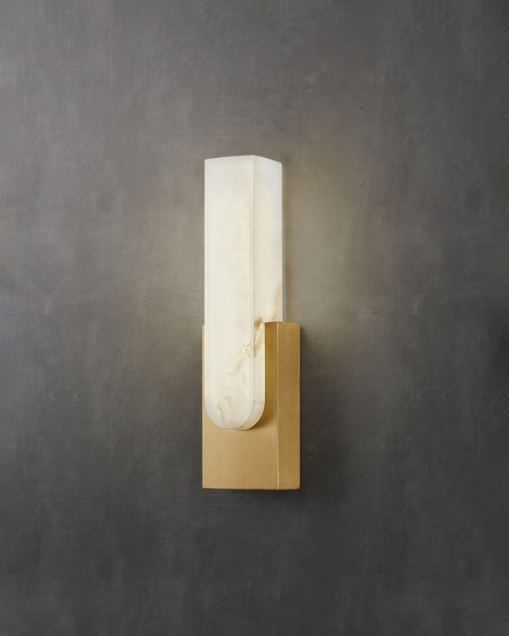 Gold-toned Clérachic wall light on a dark grey concrete-look wall, its pale alabaster bar lit softly from within.