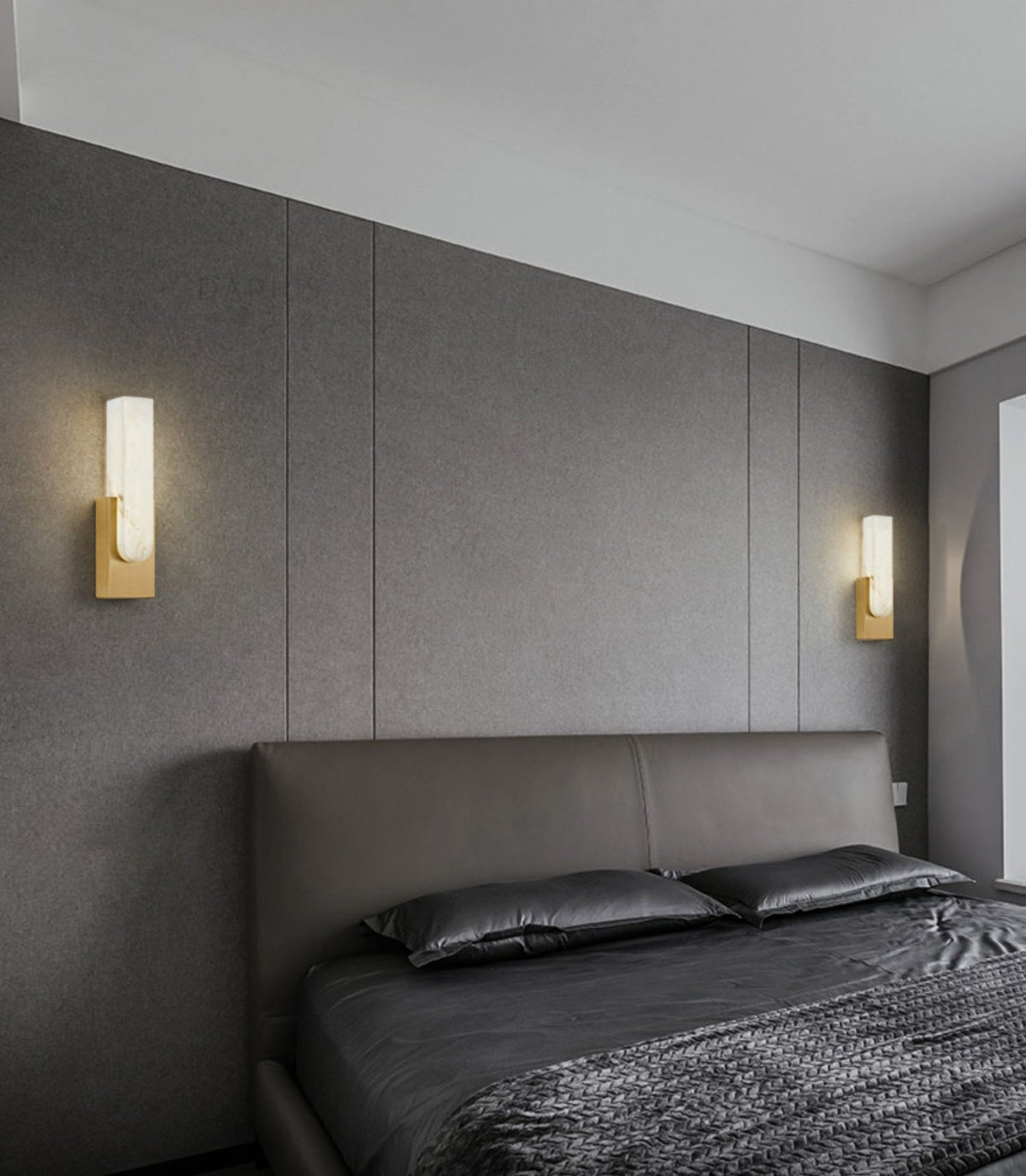 Two gold-toned Clérachic wall lights lit on either side of a grey upholstered bed, on a panelled grey wall.