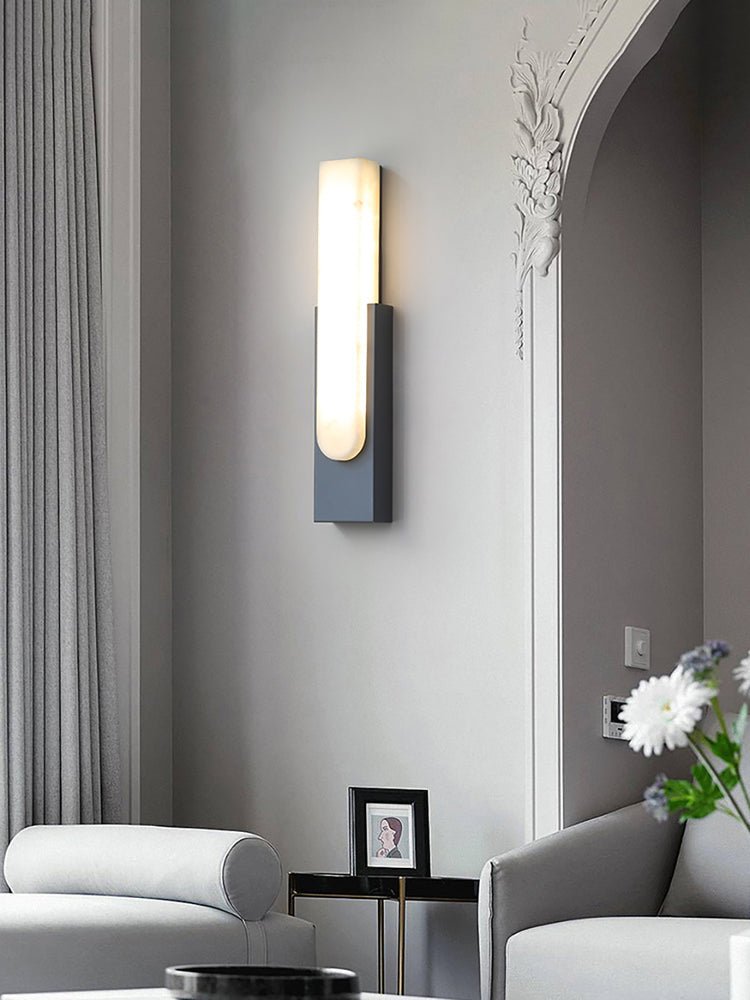 Alternative crop of the black Clérachic wall light lit on a grey wall by an arch, with grey seating and white flowers below.