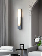 Alternative crop of the black Clérachic wall light lit on a grey wall by an arch, with grey seating and white flowers below.