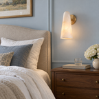 This Clérachic alabaster wall light glows beside a linen headboard, above a walnut bedside chest and a vase of white flowers.