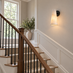 A black-backplate Clérachic alabaster sconce on a stairwell wall, glowing above white panelling and a wooden banister.