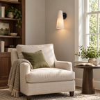 This Clérachic cone wall light with a black backplate sits above a cream armchair with a green cushion, next to a bookcase and window.