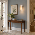A Clérachic alabaster cone sconce on a blue-grey hallway wall, framed botanical print beside it and a console table below.