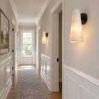 Down a long panelled hallway with a patterned runner, a Clérachic alabaster wall light with a black backplate glows in the foreground.