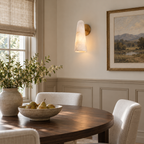 A brass-backed Clérachic alabaster sconce beside a sash window, above a round dining table set with a bowl of pears.
