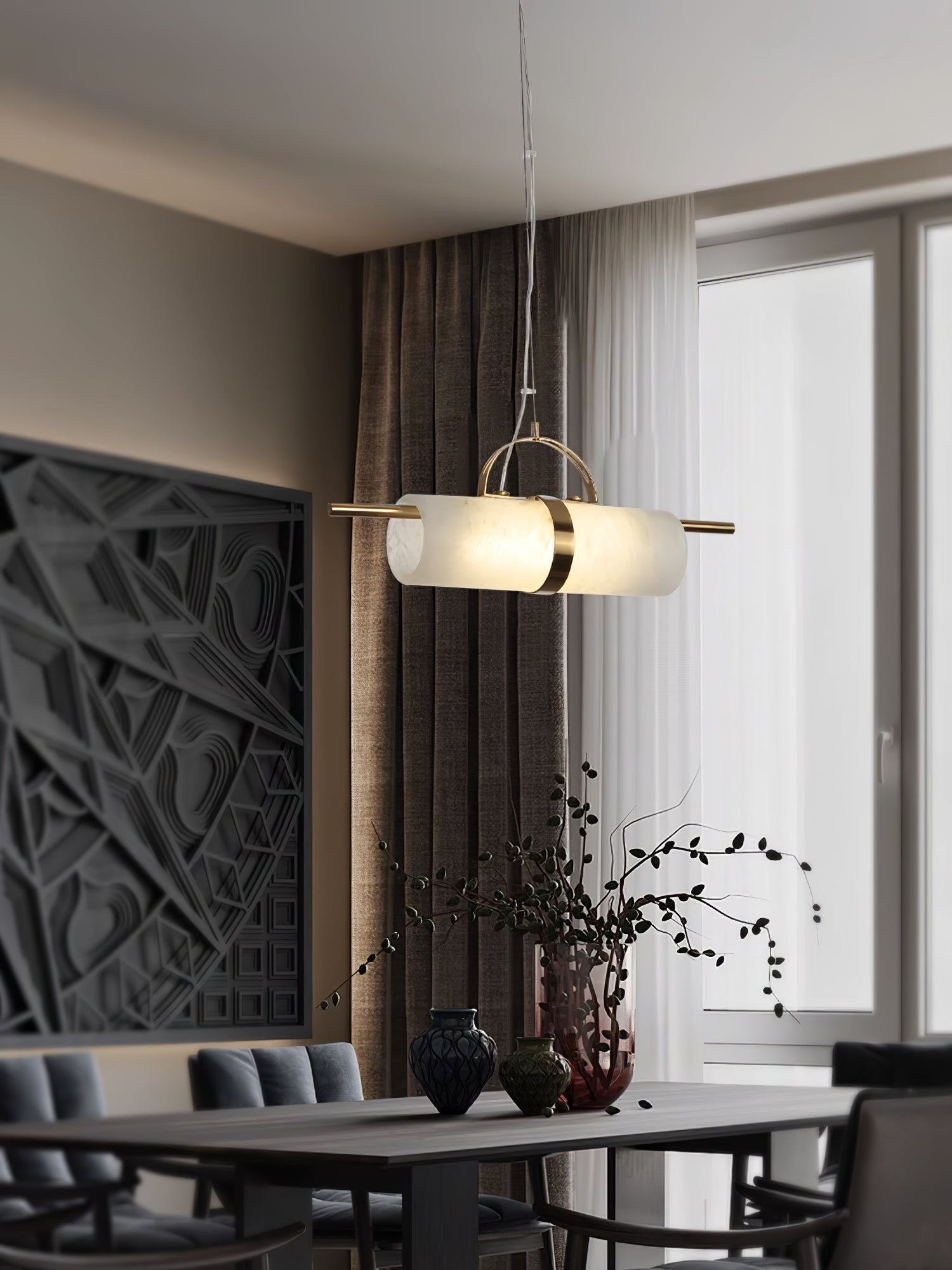 Clérachic Alabaster Cylinder LED Pendant Light glowing over a dining table by a curtained window, above dark vases and dried branches.