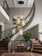 A cluster of three of these Clérachic pendants glowing amber above a curving staircase with a glass balustrade and a potted fiddle-leaf fig.