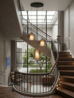 From Clérachic, three alabaster pendants lit warm in the well of a spiral staircase, with a tall black-framed window and garden behind.