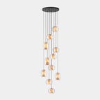 A long cluster of clear glass Clérachic pendants hangs at many heights from a wide round black canopy on a pale grey backdrop.