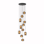 A long cluster of smoke grey glass Clérachic pendants hangs at many heights from a wide round black canopy on a white backdrop.