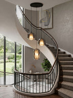 Three smoke grey glass pendants from Clérachic hang from a round black canopy beside a sweeping staircase with a dark handrail and a lawn view.