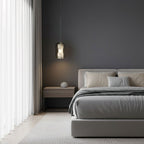 A single Clérachic alabaster pendant on a thin cord hangs against a dark grey bedroom wall, above a low bed with pale linen.