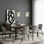 Two Clérachic alabaster pendants hang side by side over a dark wood dining table with grey chairs, in front of a black and gold artwork.