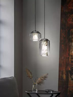 Two Clérachic pendants at different heights against a grey wall, the larger one showing grey-veined alabaster, with dried grasses on a side table below.