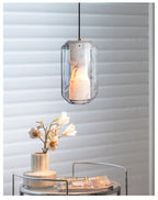 A Clérachic alabaster pendant hangs in front of white slatted blinds, above a glass side table holding a vase of blossom and a magazine.
