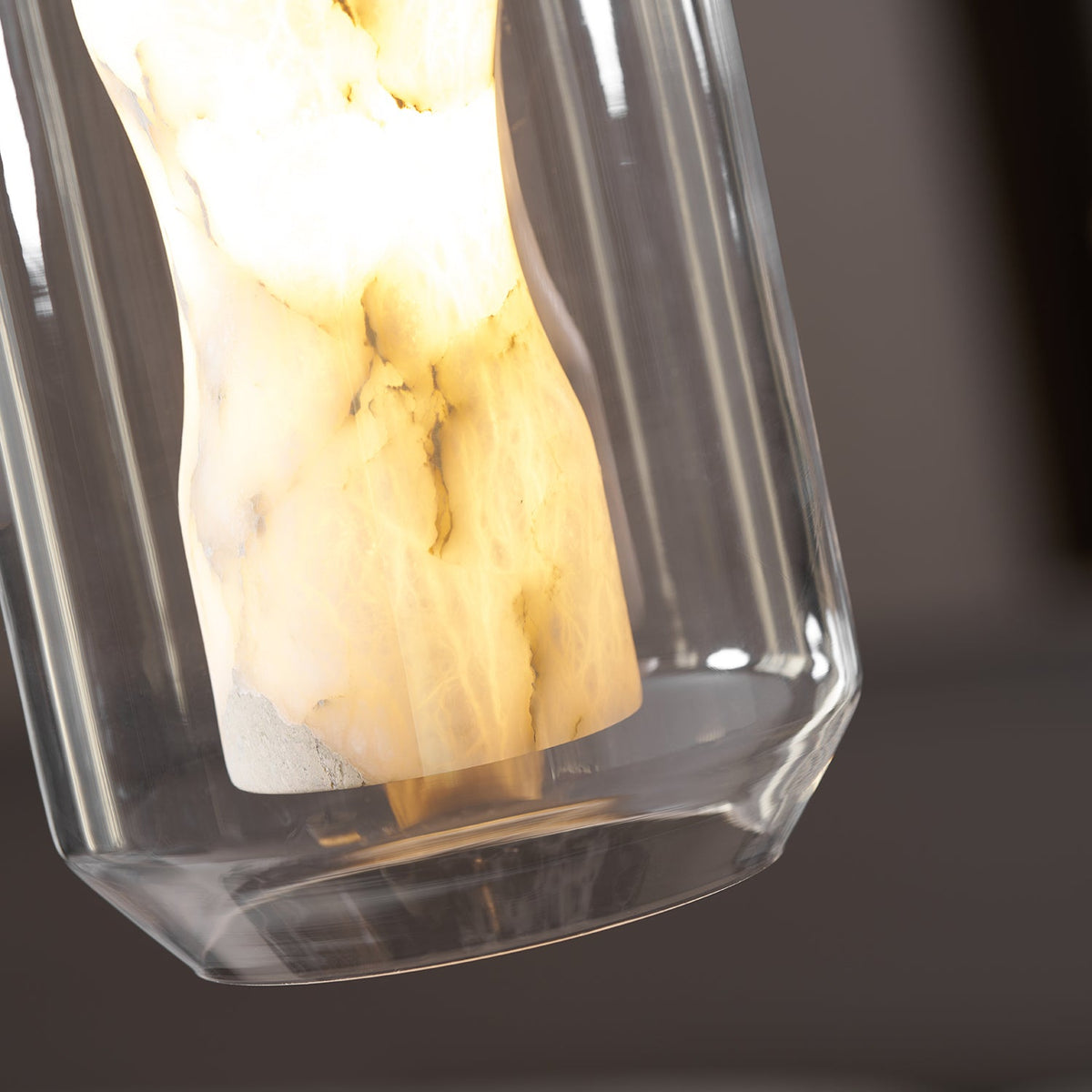 Close view of the lit alabaster column and the clear glass rim at the base of this Clérachic pendant, against a dark blurred background.