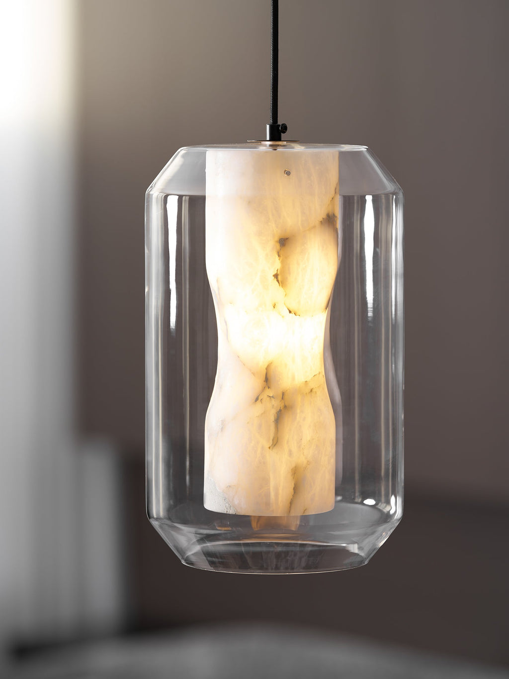 A tall, faceted-glass Clérachic pendant with a lit alabaster column, hanging on a black cord in front of a soft grey interior.