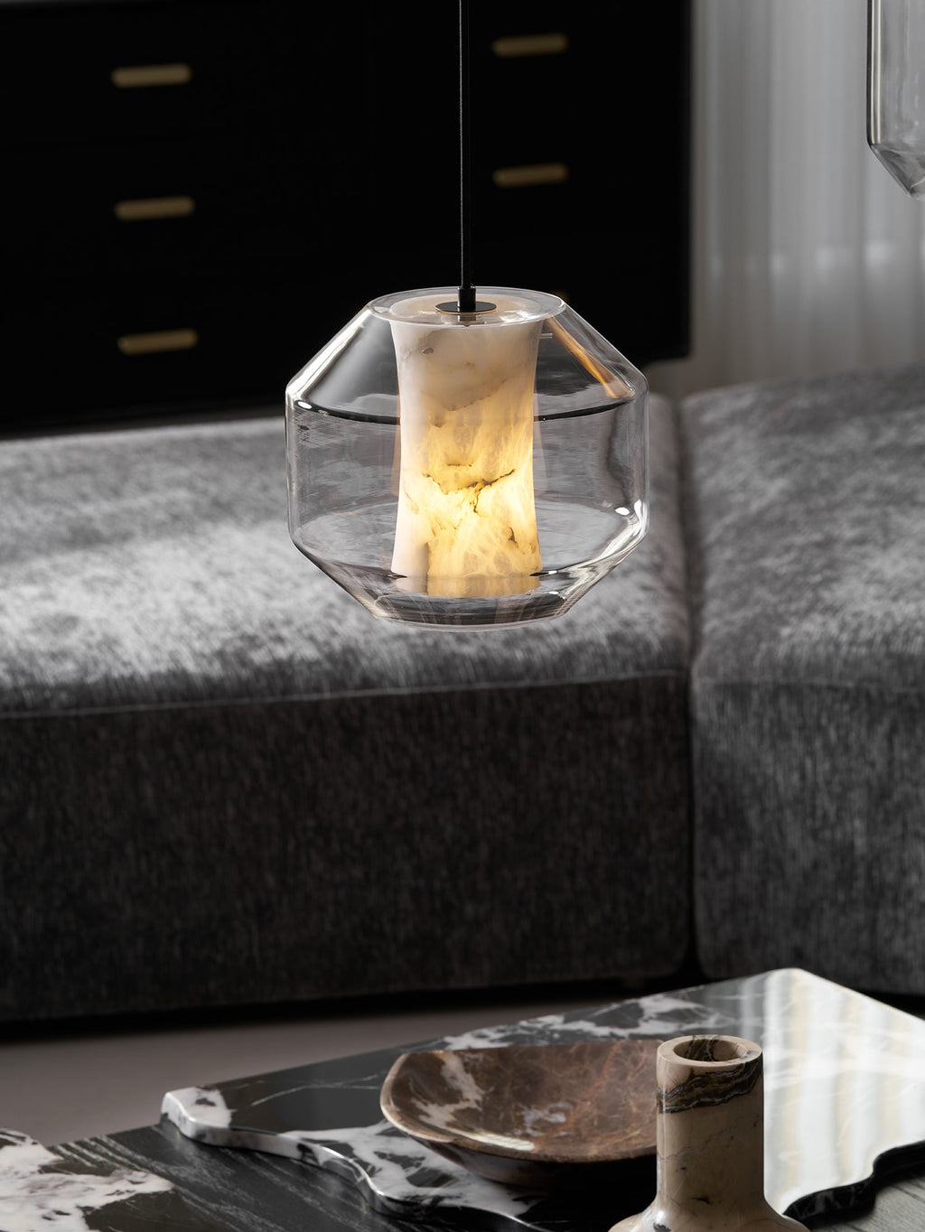 A wide faceted-glass Clérachic pendant glows warm above a grey upholstered sofa, with a dark marble table and a brown dish below.