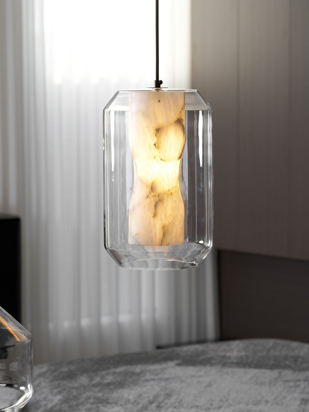 A tall faceted-glass Clérachic pendant with a lit alabaster column, hanging in front of sheer curtains and grey seating.