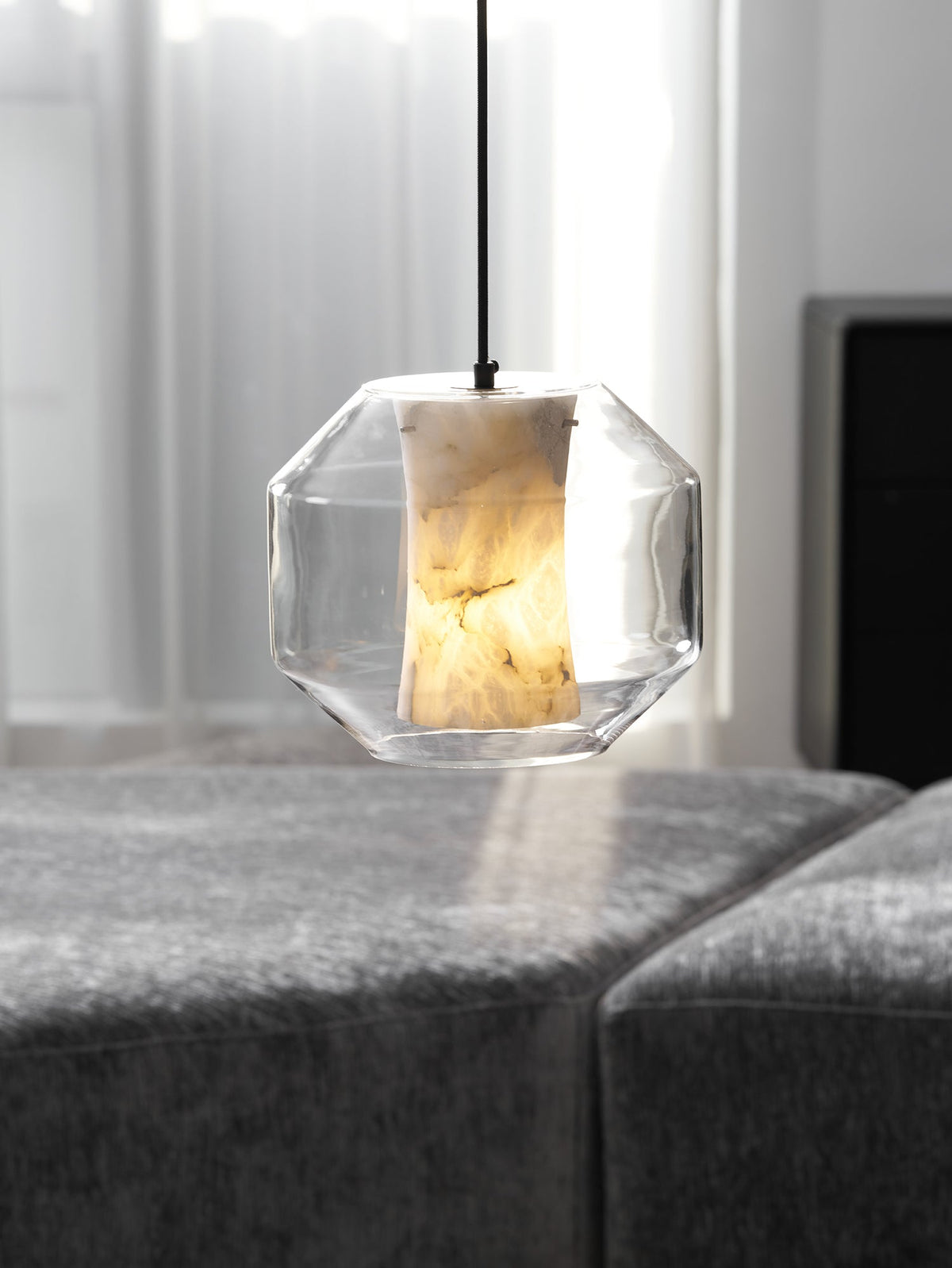 A wide, faceted-glass Clérachic pendant lit warm above a grey ottoman, with sheer curtains softening the window behind.