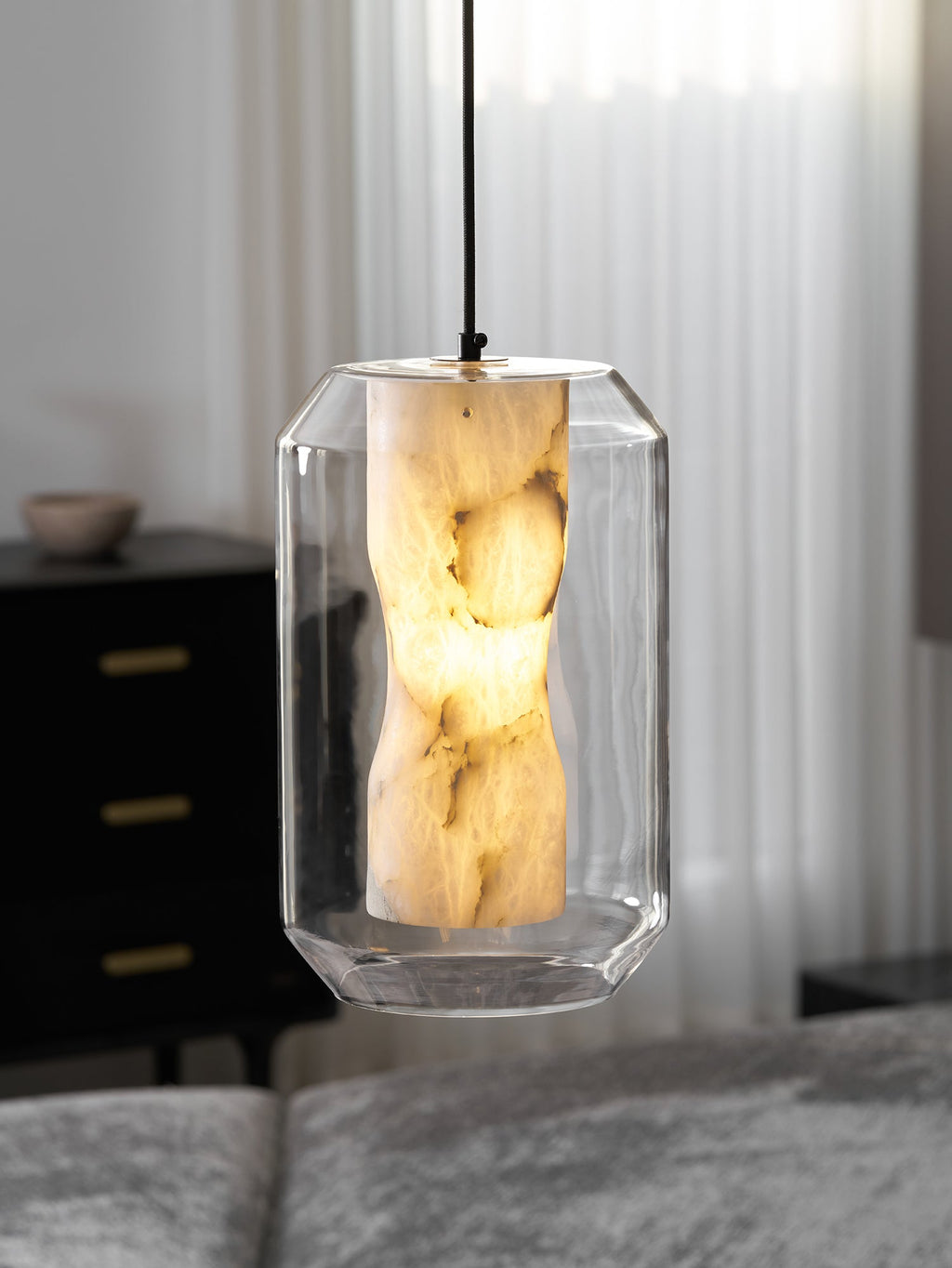 A tall faceted-glass Clérachic pendant glowing warm in front of sheer curtains, with a dark chest of drawers blurred behind.