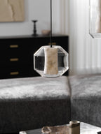 A wide Clérachic pendant with an unlit alabaster column hangs above a grey sofa, with a dark cabinet and a pale artwork behind it.