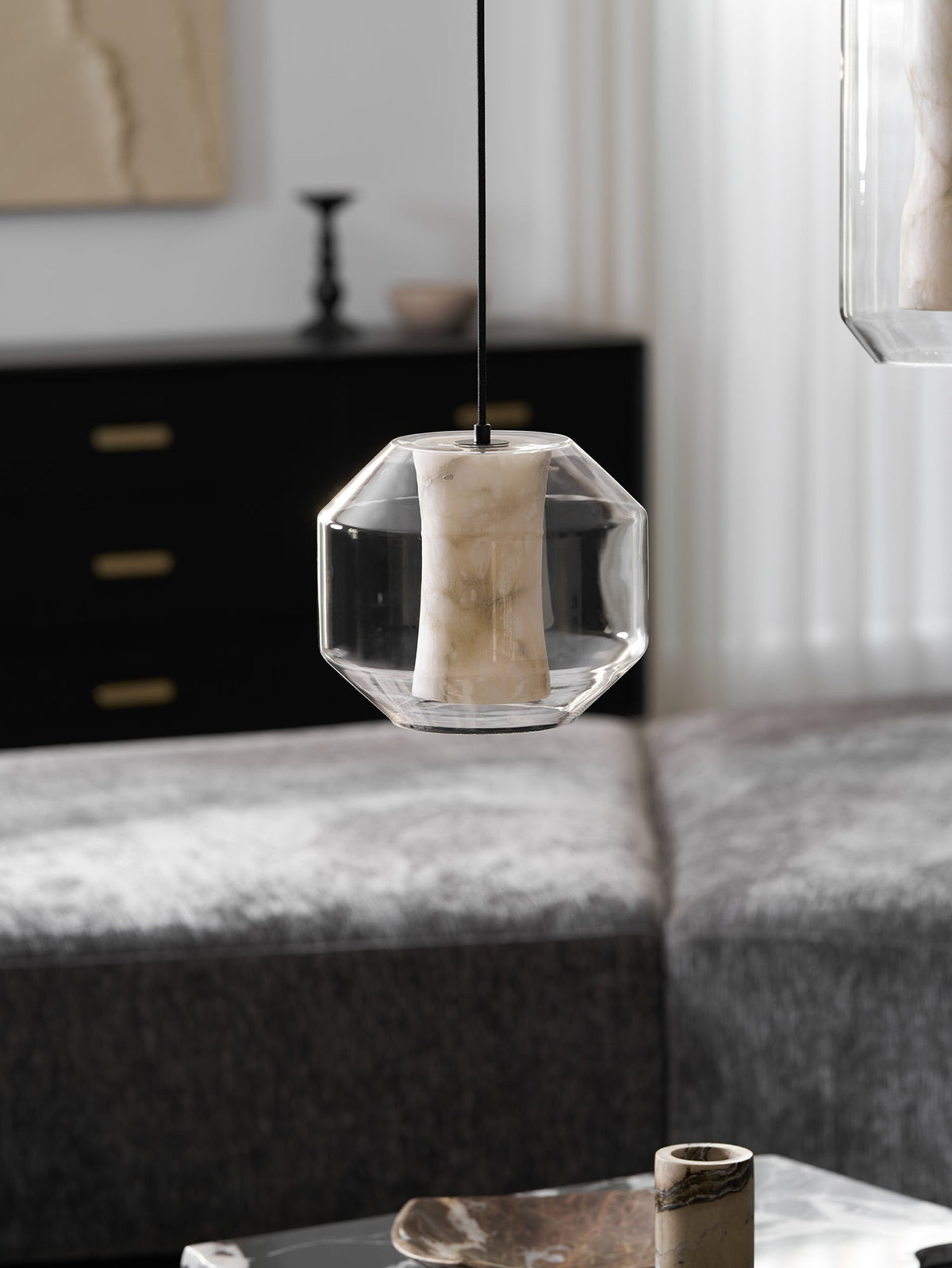 A wide Clérachic pendant with an unlit alabaster column hangs above a grey sofa, with a dark cabinet and a pale artwork behind it.