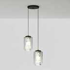 Two tall clear glass Clérachic pendants with veined alabaster hang at different drops from a round black canopy against a pale grey wall.