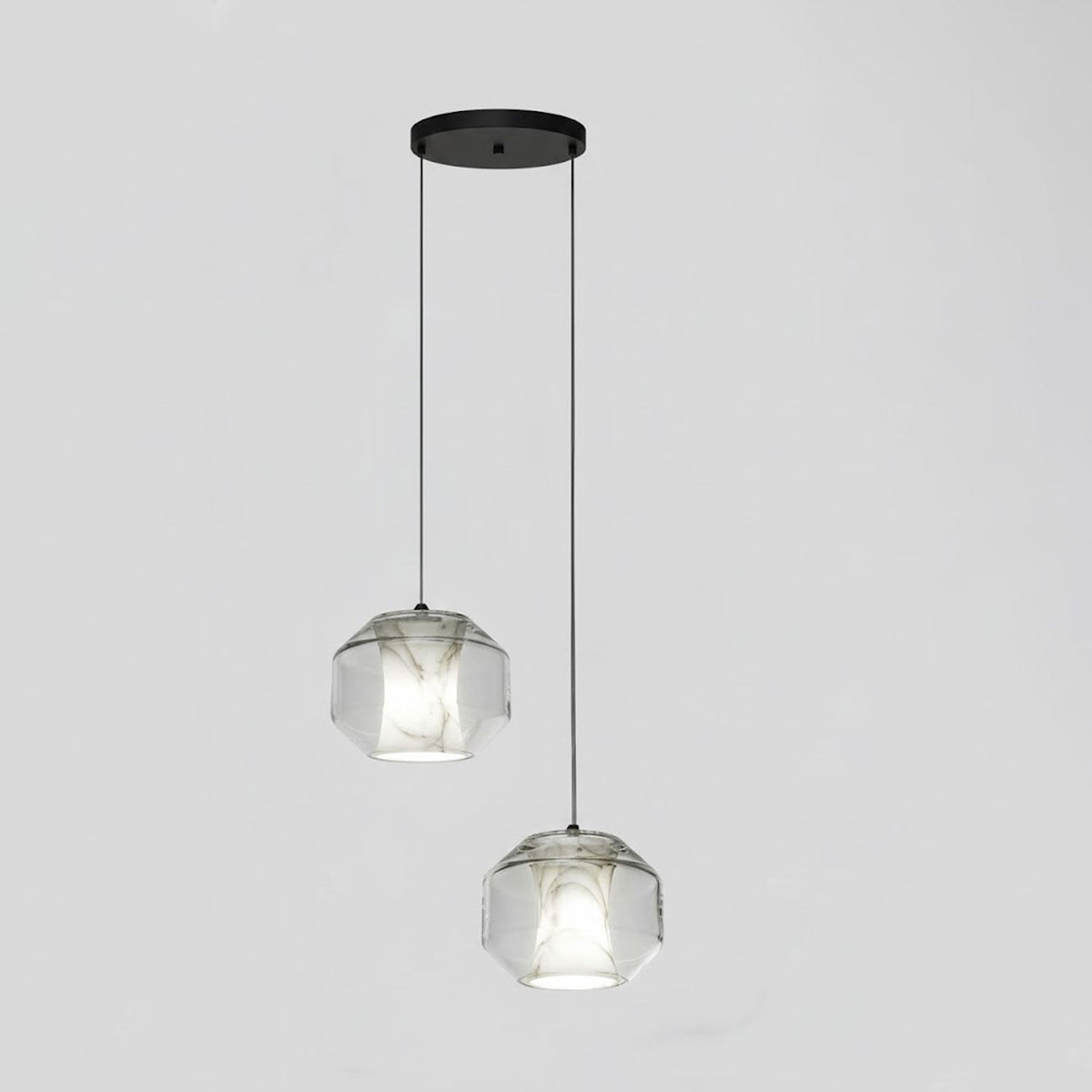 Two wide clear glass Clérachic pendants with white alabaster hang at different heights from a black canopy against a pale grey wall.