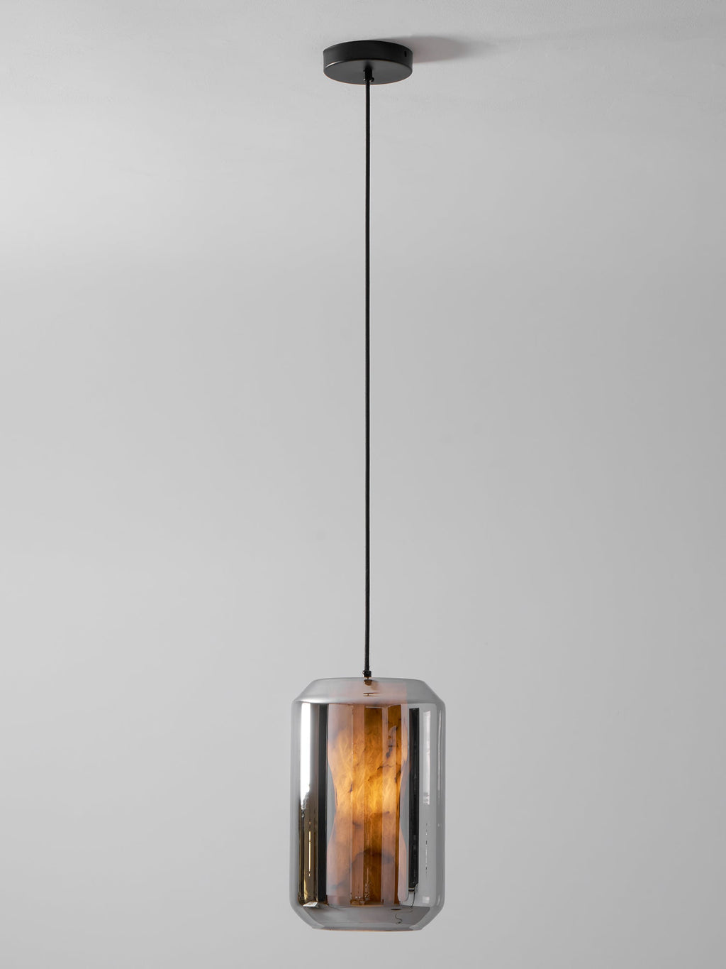 A tall smoke grey glass Clérachic pendant on a black cord and canopy, its alabaster column glowing amber against a pale grey wall.